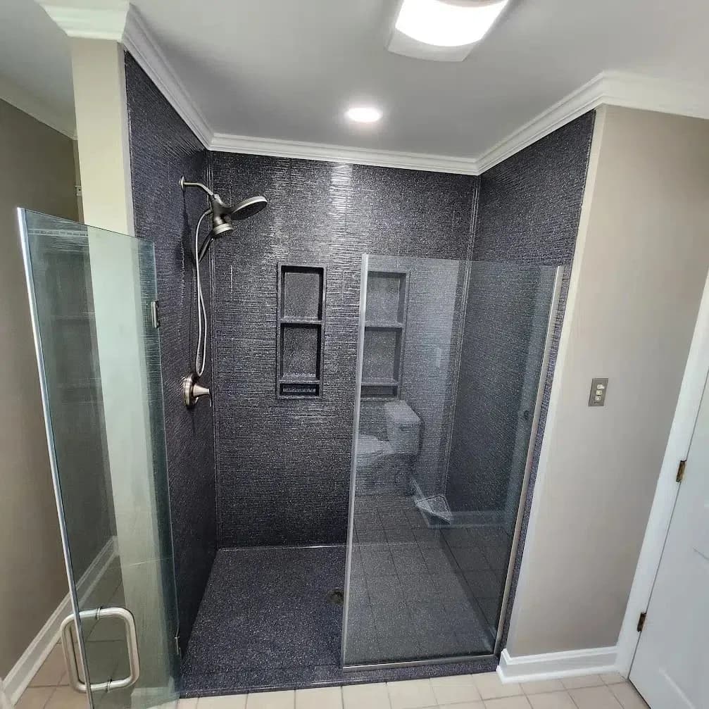 A new walk-in shower in an NW Arkansas home with dark blue surround, glass doors, seating, plenty of storage space in the back walls, and modern hardware, including a hand-held showerhead.
