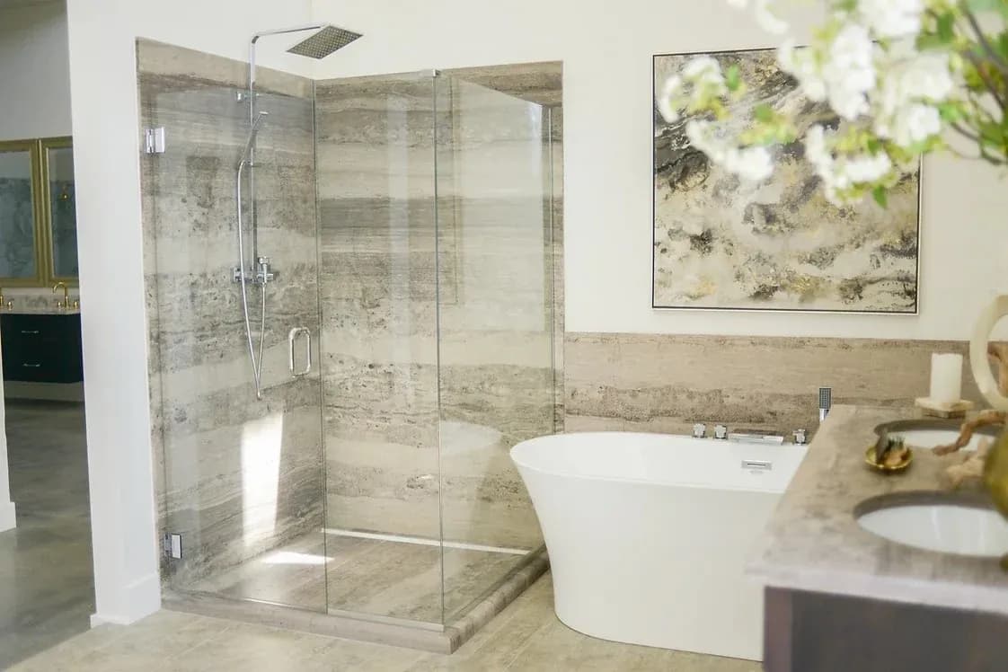 A furnished wet area of a residential bathroom with a new walk-in shower with new hardware, glass walls, a door, and a soft brown stone surround.